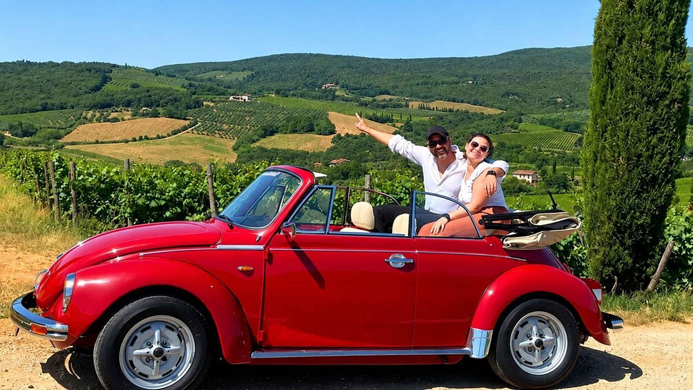 A red vintage convertible car parked on a dirt road with a scenic vineyard and rolling hills in the background. 1