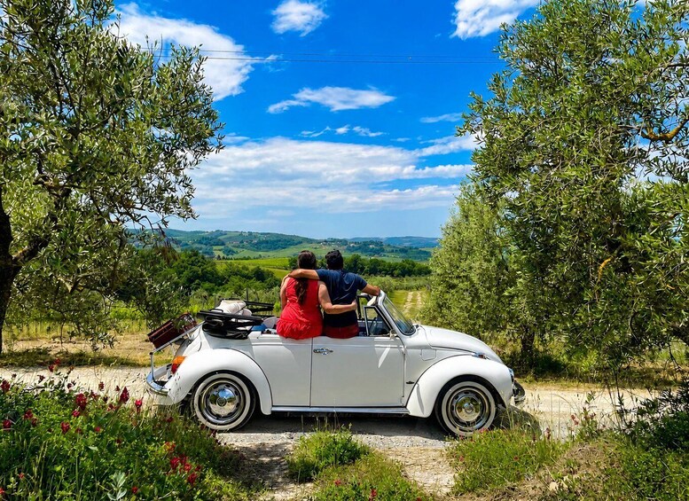 A white convertible car parked on a rural road with a couple embracing, surrounded by olive trees and a scenic countryside view. 5