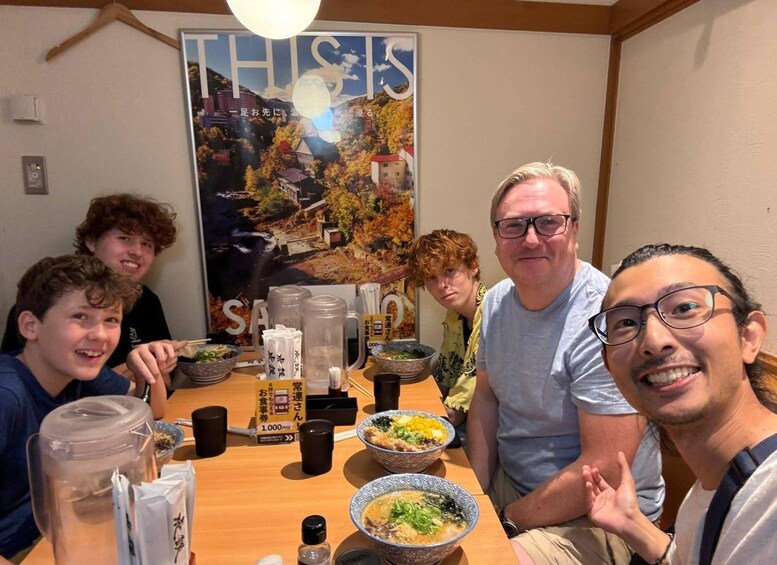 A group of people sitting around a table with bowls of food, a poster on the wall, and a wooden ceiling. 3