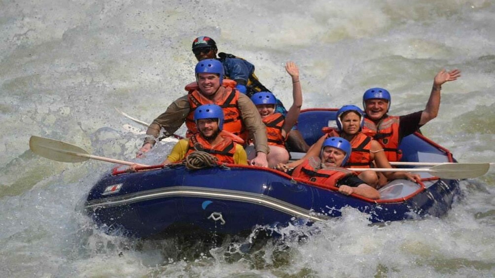 A group of people in a blue raft with paddles, wearing life jackets and helmets, navigating through choppy water. 3
