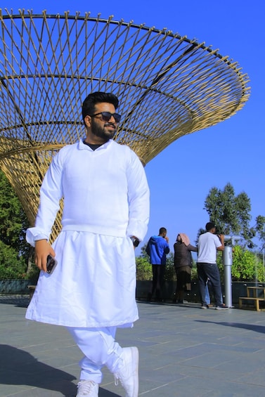 A person in white attire walking in front of a large, geometric structure with a clear blue sky in the background. 2