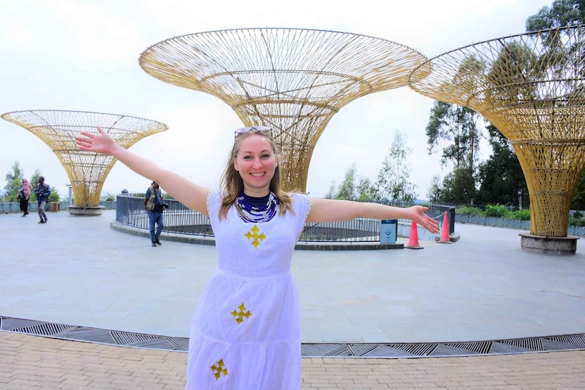 A person in a white dress with yellow stars, standing in a public space with large, circular, woven structures in the background. 1