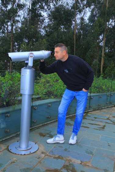 A man using a telescope on a platform with a metal railing, surrounded by greenery. 4