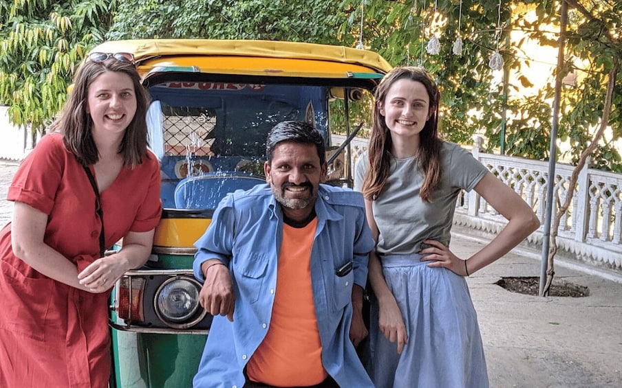 Three people posing with an auto-rickshaw in a garden-like setting with trees and a white fence. 4