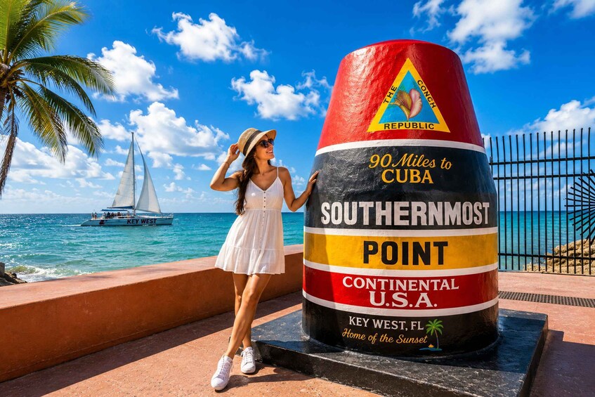 A woman near a large sign at Key West, FL, with a sailboat in the background. 2