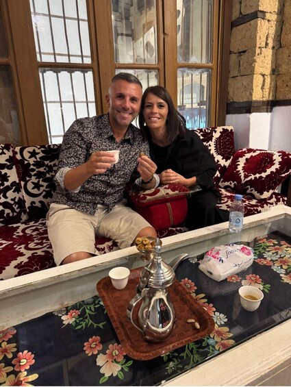 A man and a woman sitting on a patterned sofa, holding a small cup and a bowl, with a tray of food and a teapot on a table. 3