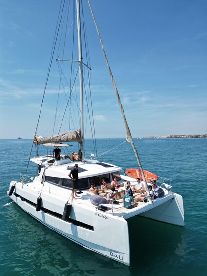 A white catamaran with the name 'BALI' on its side, sailing on clear blue water with a cityscape in the background. 1