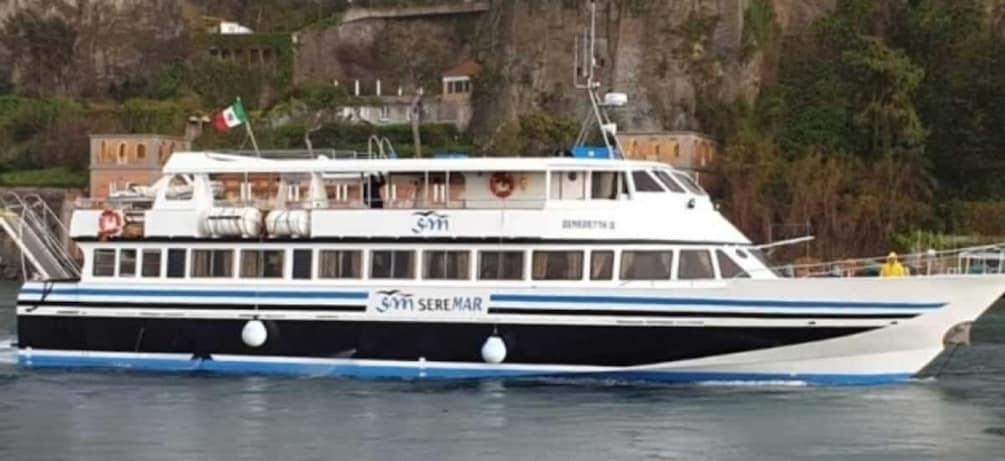 A boat named 'S.M. SEREMAR' with a flag, docked near a rocky coastline with buildings in the background. 2