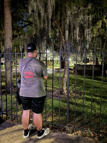 A man standing at a cemetery gate, wearing a t-shirt with a slogan and black shorts. 5