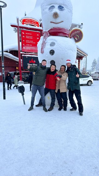 Un groupe de personnes pose devant une grande sculpture de bonhomme de neige, avec une pancarte indiquant le nom du « Village des fêtes du Père Noël ». 1