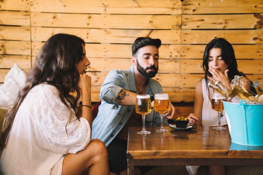 Three people sitting at a wooden table with beer glasses and a bucket of olives. 2