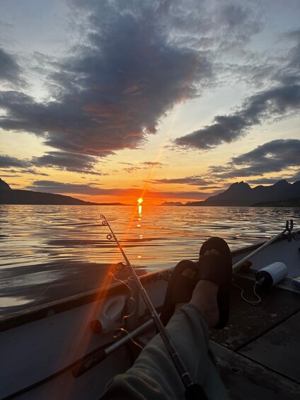 A person fishing at sunset with a boat on calm water, mountains in the background, and a cloudy sky. 5