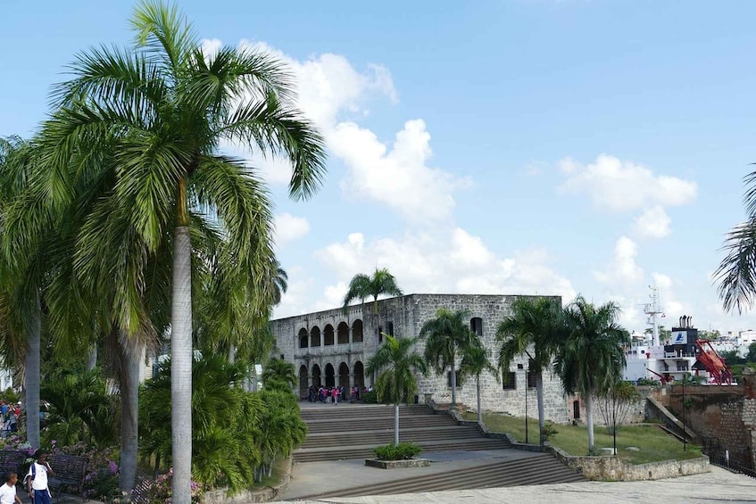 A historic stone building with arched windows and a staircase leading up to it, surrounded by palm trees and a clear sky. 5