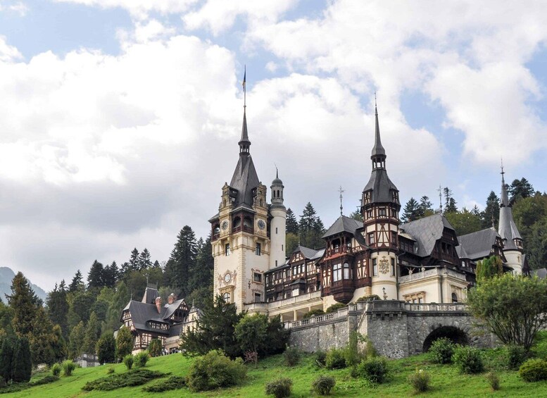 A historic castle with multiple towers and a clock tower, surrounded by lush greenery and mountains. 4