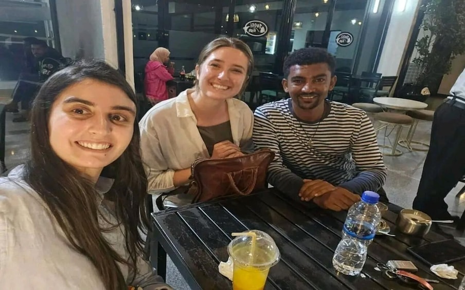 Three people seated at an outdoor cafe with a table, a drink, and a water bottle. 2