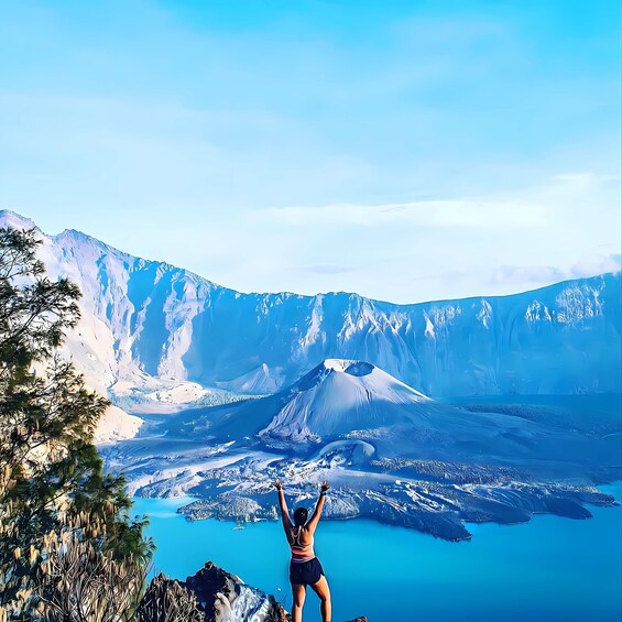 A person standing on a mountain peak with arms raised, overlooking a valley with a lake and snow-capped mountains. 5