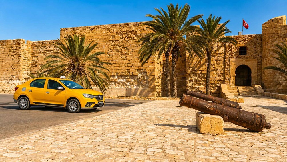 A yellow car parked in front of a stone wall with palm trees and a flag. 1