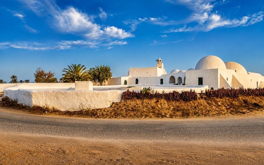 A white building with domed structures and a bell tower, surrounded by dry vegetation and palm trees. 2