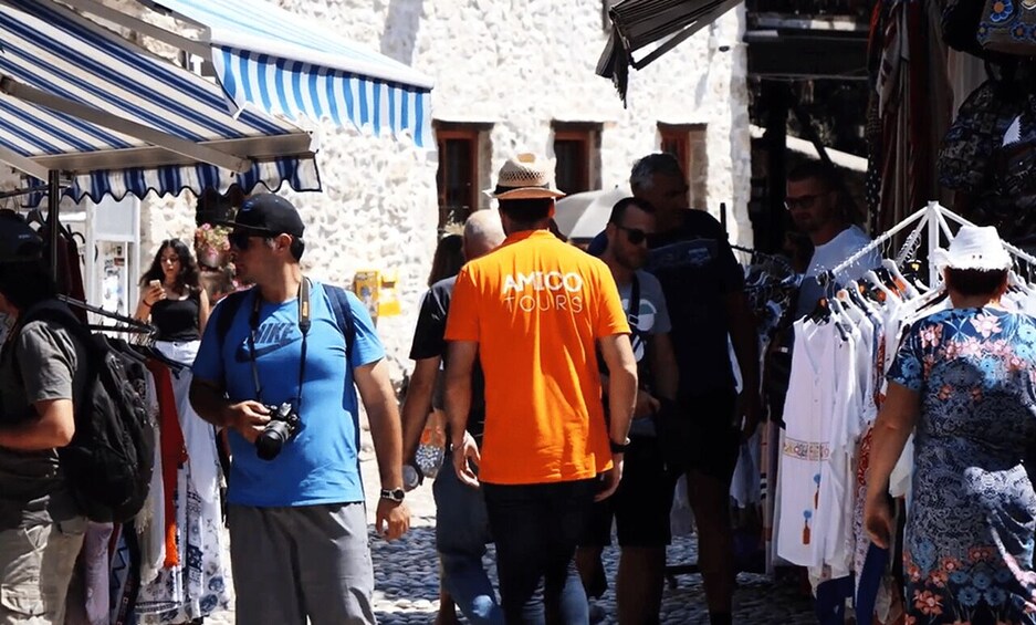 A crowded street with people walking, a man in an orange shirt, and a stall with clothes. 2