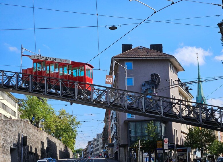 A red tram labeled 'POLYBAHN' on a track, a building with the number '48' on its facade, and a green spire in the background. 3