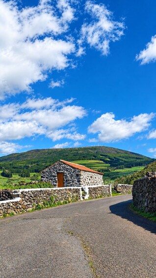 A stone building with a red roof, a stone wall, and a road leading to a hill with a green landscape. 3