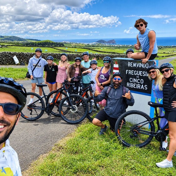 A group of cyclists posing in front of a sign with a scenic view of the countryside and the ocean in the background. 4