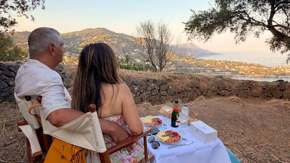 A couple sitting at a table with a scenic view of a coastal town and mountains in the background. 5