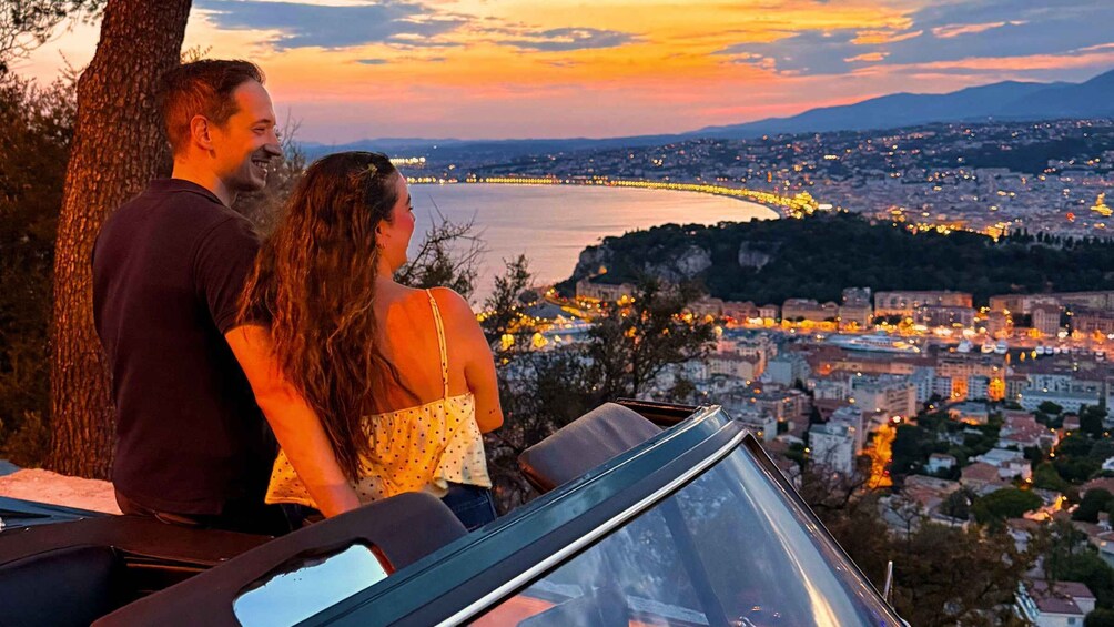 A couple overlooking a cityscape at sunset with a view of a body of water and mountains in the distance. 1