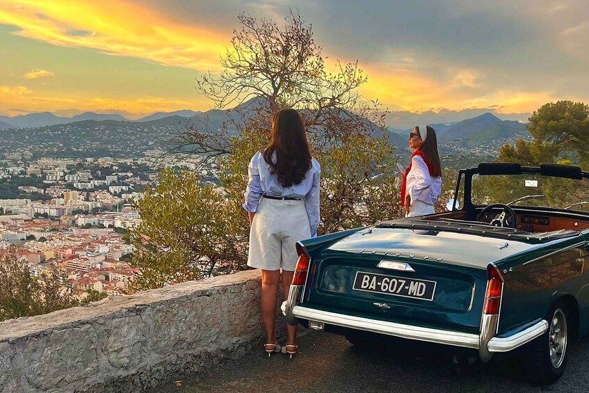 A woman standing next to a green convertible car with a license plate BA-607-MD, overlooking a cityscape with mountains in the background. 1