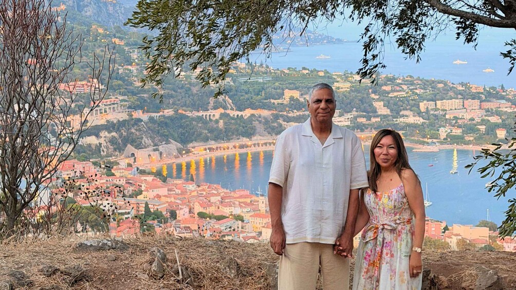 A man and a woman standing on a hill overlooking a coastal town with a bridge and buildings. 3