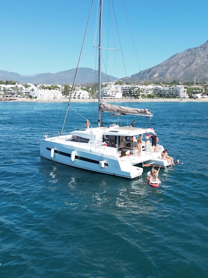 A white catamaran with people on board, sailing on a calm sea with a coastal town and mountains in the background. 4