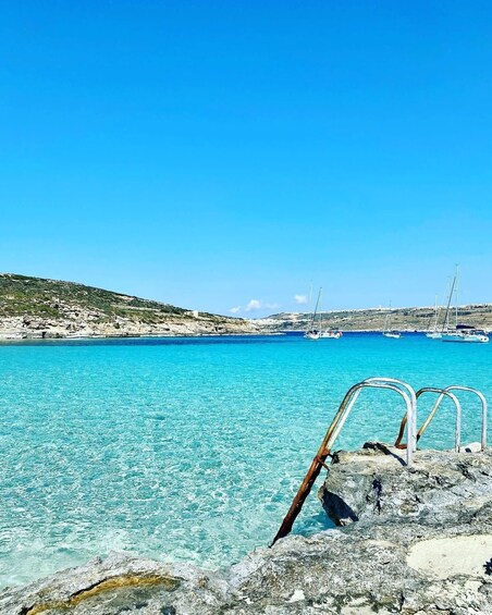 A coastal scene with clear turquoise water, rocky shore, and sailboats in the distance. 1