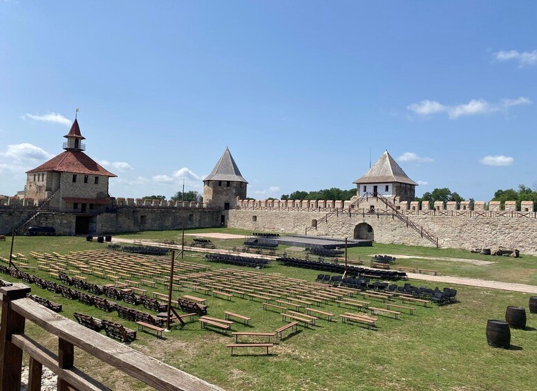 A historic fort with stone walls and two towers, a grassy courtyard with wooden benches, and a clear blue sky. 5