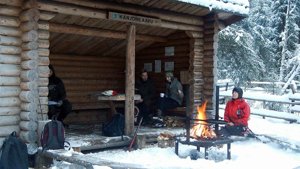 A wooden cabin with a sign reading "KANJONILAAVU". Inside, people are seated around a table with a fire burning outside. 4