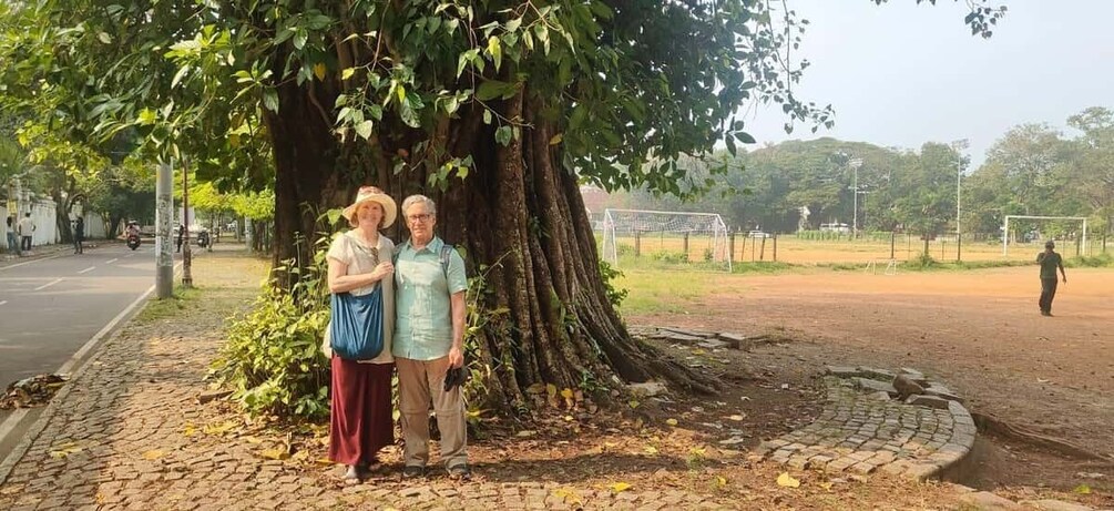 Two people standing under a large tree on a paved path with a soccer field in the background. 2