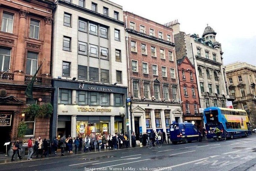 Une rue où une file de personnes attend, un autobus et des immeubles abritant des commerces tels qu’un magasin Tesco et une banque. 1