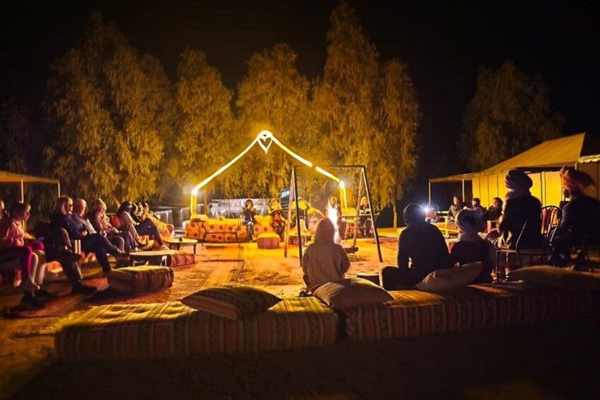 An outdoor gathering with people seated on cushions, a stage with a tent, and trees in the background. 1