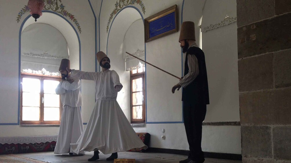 A person in traditional attire holding a staff, standing in a room with arched windows and a framed certificate on the wall. 5
