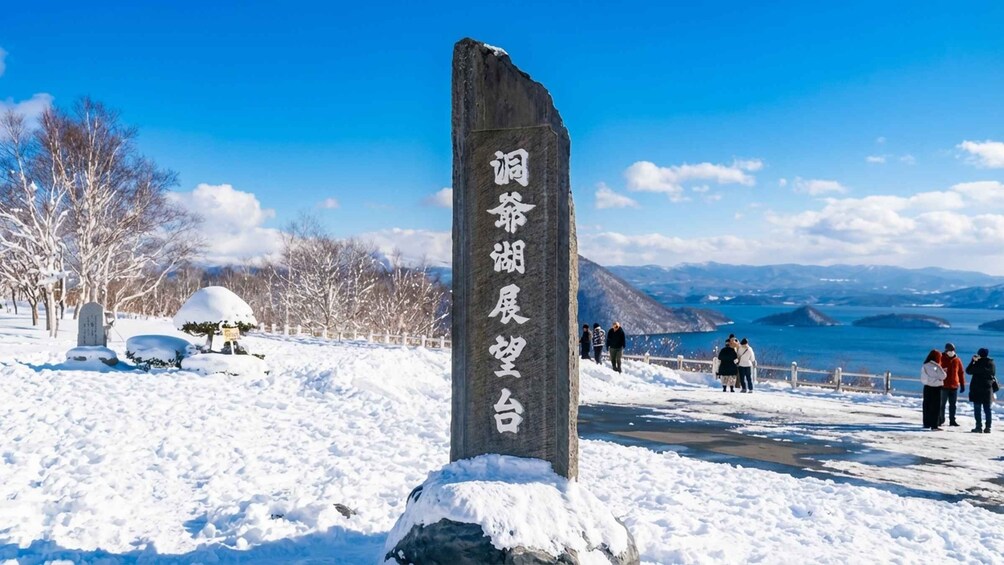 A snow-covered landscape with a stone monument and a view of mountains and a lake. 5