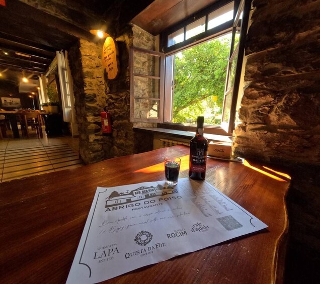 A wooden table with a bottle of wine and a menu at a restaurant with stone walls and a window. 2