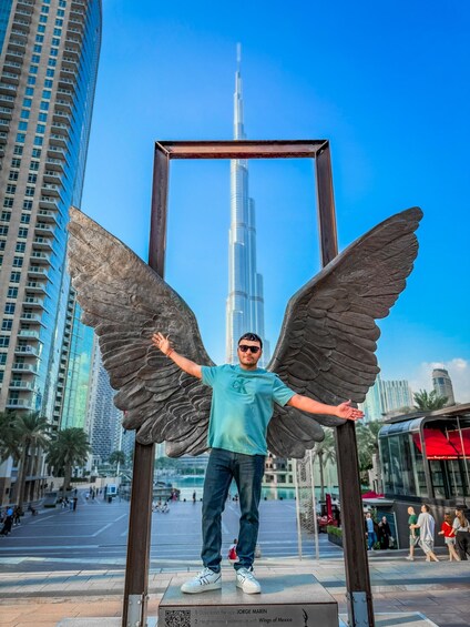 A person standing in front of a large winged sculpture with a tall building in the background. 3