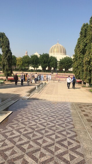 A public square with a patterned tile floor, a large dome structure in the background, and people walking around. 3