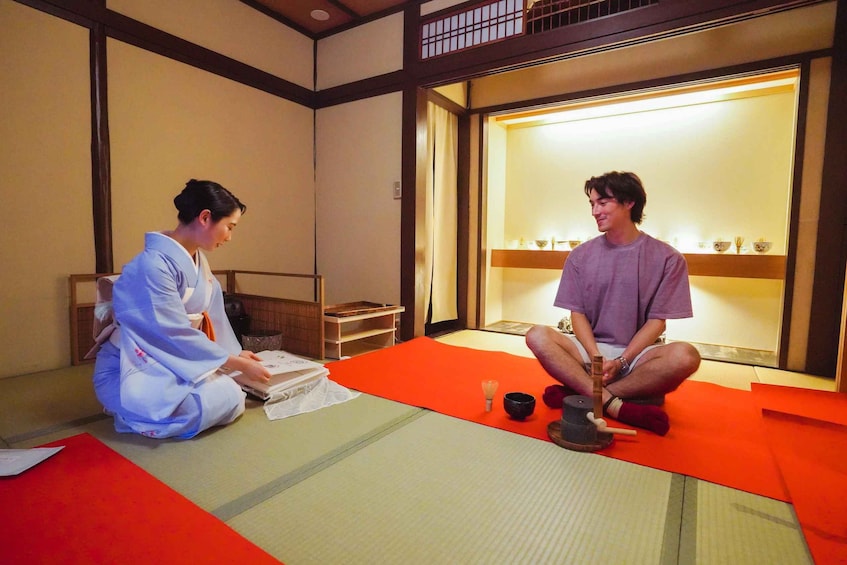 Two people sitting on tatami mats in a traditional Japanese room with sliding doors and a shelf with bowls. 1