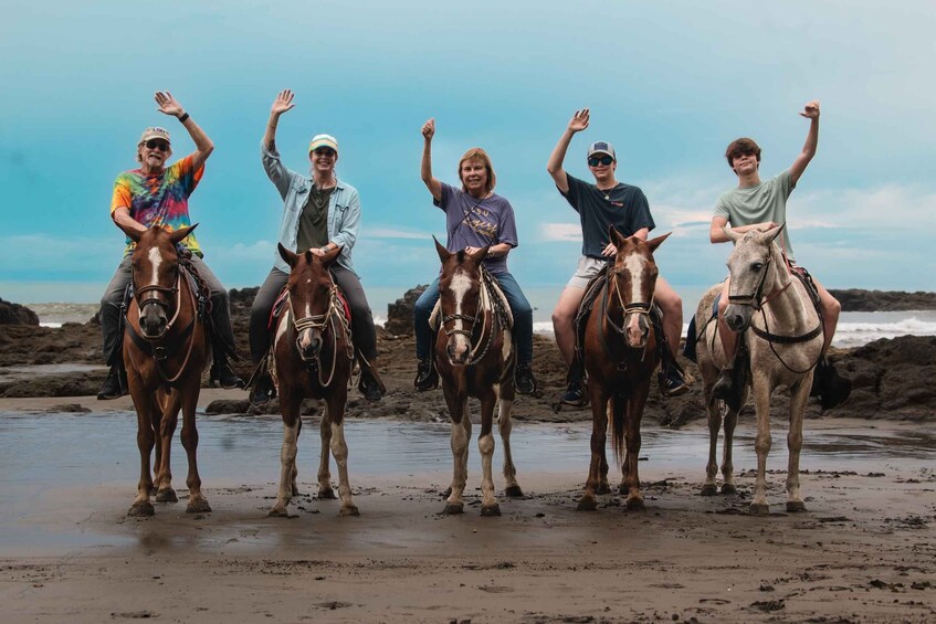 Five people on horses on a beach with waves in the background. 3