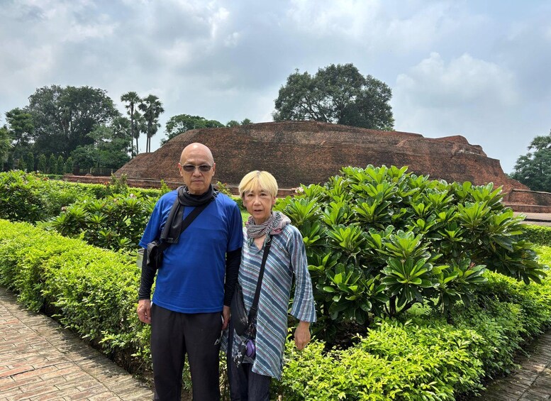Two people standing in front of a large brick structure with lush green bushes and trees in the background. 2