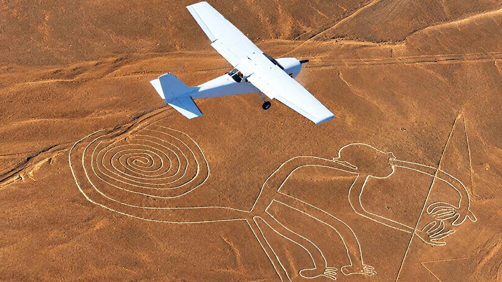 A white airplane flying over a desert landscape with large spiral and human figures etched into the ground. 1