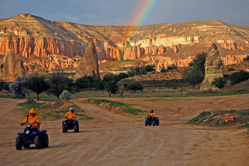 Three people riding ATVs on a dirt road with a canyon in the background and a rainbow. 1