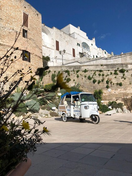 A white tuk-tuk parked in front of a stone wall with greenery. 1