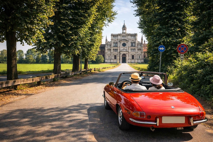 A red convertible driving on a tree-lined road towards a historic building. 1