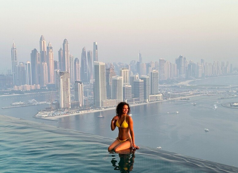 A woman in a yellow bikini sitting on the edge of an infinity pool overlooking a city skyline. 1